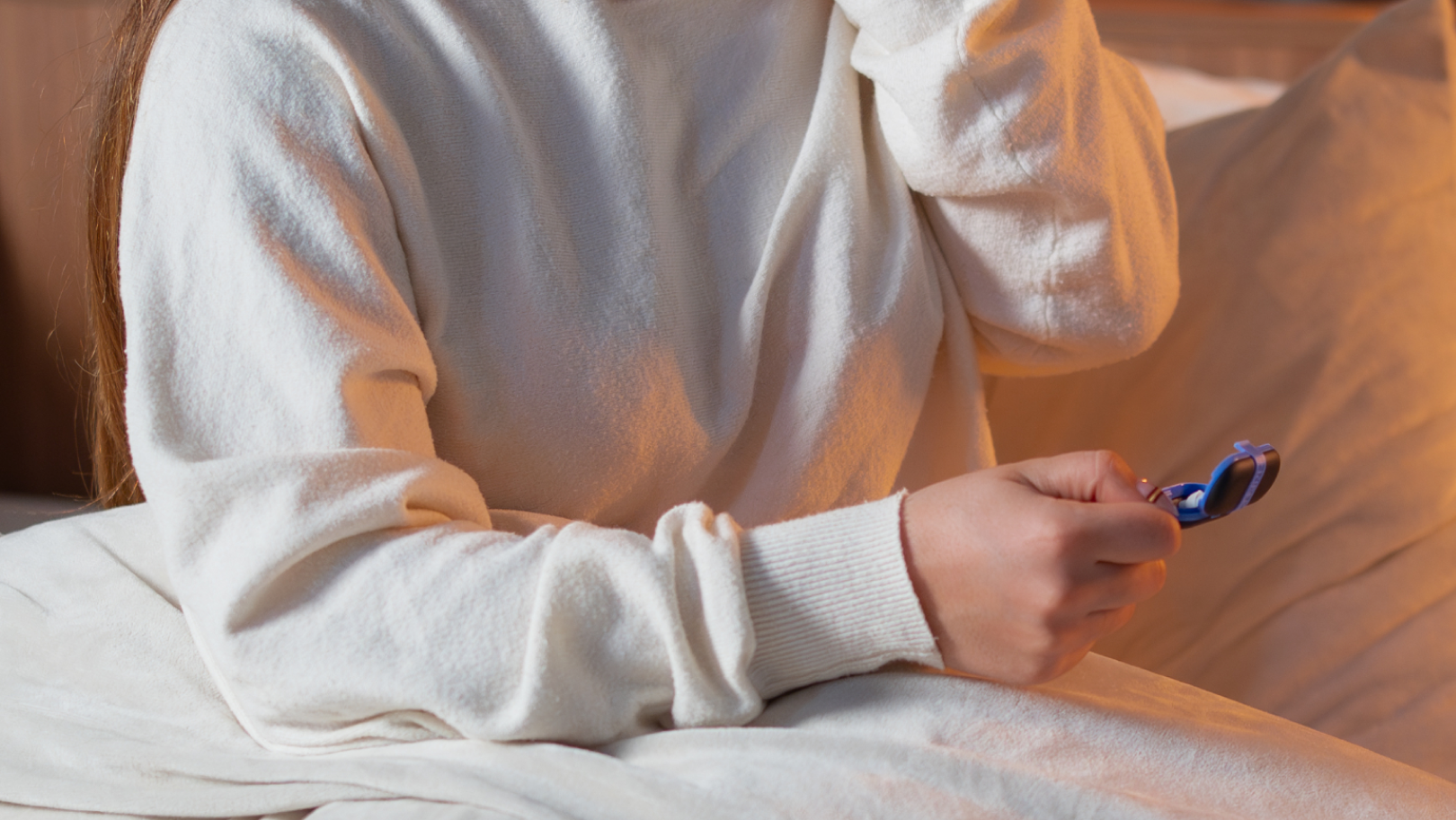 Woman wearing a beige sleep mask in a cozy bedroom setting putting in sleep earplugs from alpine